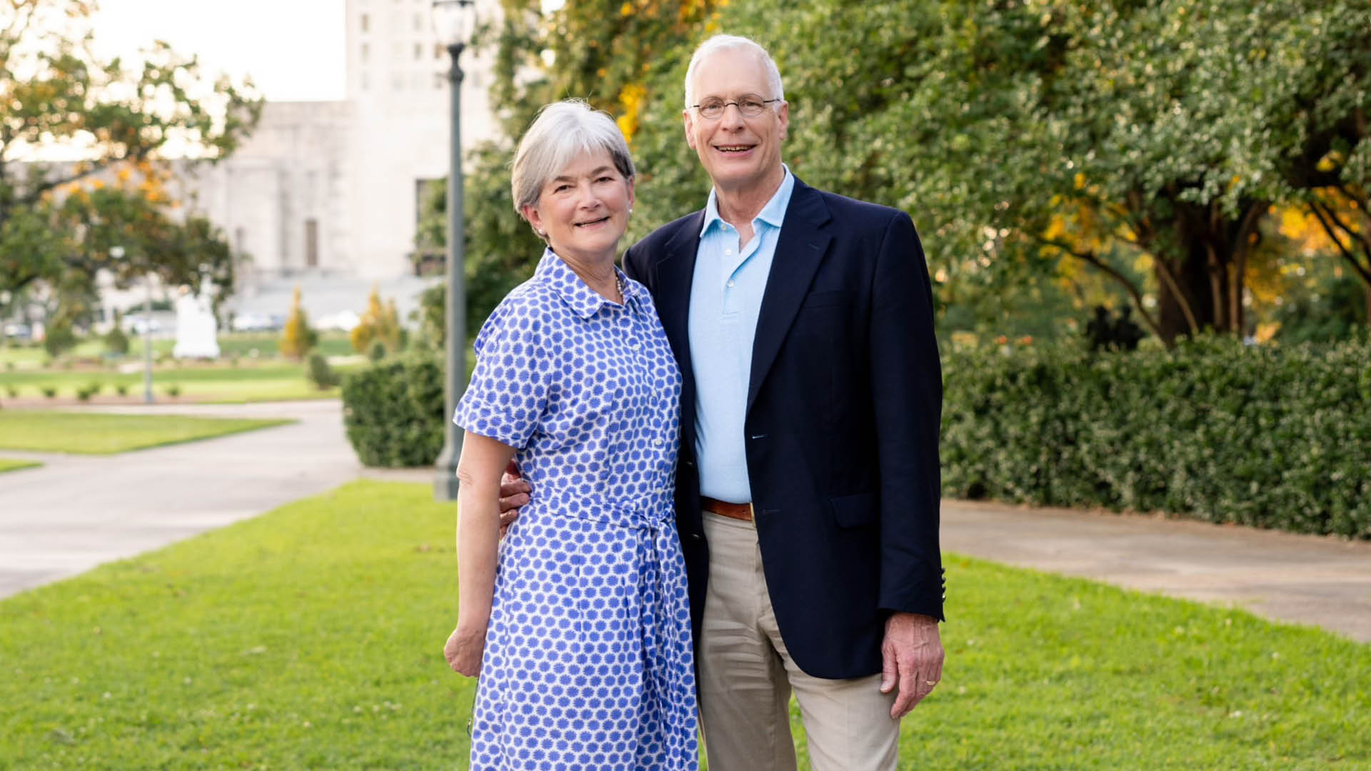 An older woman and man stand together outdoors on grass, facing the camera and smiling. The woman is wearing a blue patterned dress. The man is wearing glasses, a dark blazer, a light blue shirt, and khaki pants. There are trees, bushes, a sidewalk, a lamppost, and a light-colored building in the background